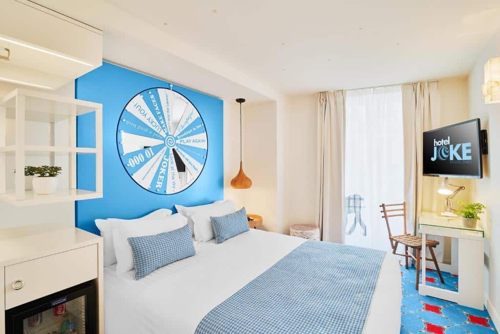 A imagem mostra um quarto vibrante e moderno no Hotel Joke - Astotel, um dos hotéis com café da manhã em Paris. A cama é grande, com lençóis brancos e almofadas azuis, situada contra uma parede azul com uma grande roda da sorte decorativa. Há uma janela alta com cortinas claras, permitindo a entrada de luz natural. Na parede oposta à cama, há uma televisão montada com a logo "Hotel Joke". Um pequeno frigobar está ao lado da cama, e uma mesa de vidro com uma cadeira de madeira está próxima à janela. O quarto tem um carpete azul com padrões vermelhos, criando um ambiente alegre e acolhedor.