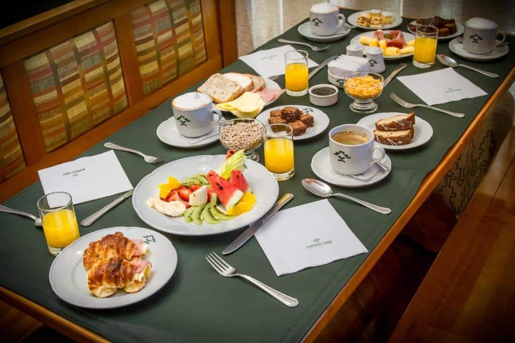 Café da manhã do Hotel Kosten Aike. Uma mesa com pratos com frutas, pães, bolos, frios, sucos, cafés e talheres.