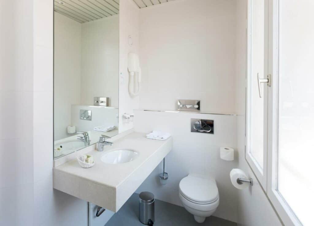 Banheiro do Hôtel la Residence Du Berry. Na parede esquerda está a pia com amenidades de banho, secador e espelho. No centro fica a privada e a lixeira, e ao lado direito está a janela.