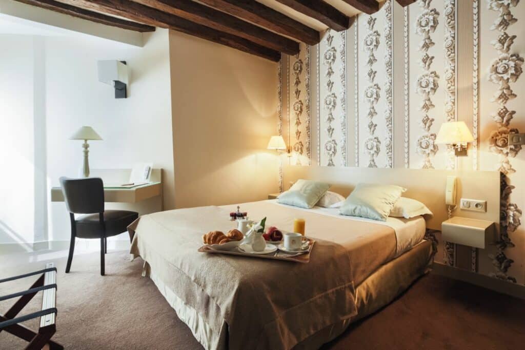 Quarto do Hôtel la Residence Du Berry, um dos hotéis perto do Palácio de Versalhes. Uma cama de casal está encostada na parede ao lado direito e tem mesinhas de cabeceira e abajures dos dois lados, assim como uma bandeja de café da manhã aos pés. Ao lado esquerdo do quarto há uma mesinha com cadeira e abajur. O teto tem ripas de madeira decorativas.