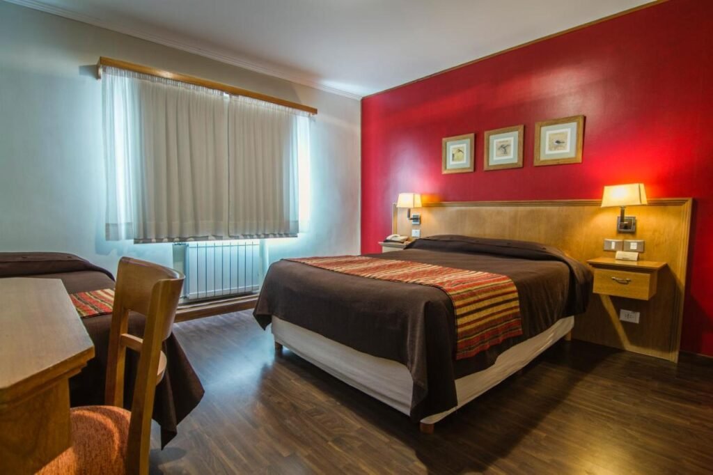 Quarto do Hotel Milan. Uma cama de casal do lado direito, de cada lado um abajur. Do lado esquerdo uma mesa de trabalho e uma cama de solteiro.