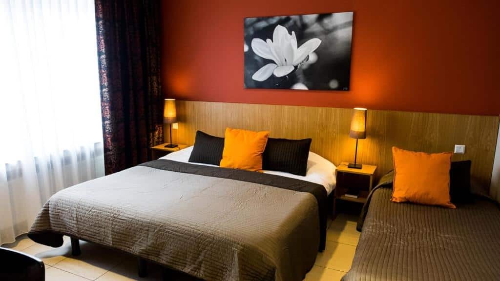 Quarto do Hotel Pax, para ilustrar o post sobre hotéis em Luxemburgo. Há uma cama de casal no centro, e outra de solteiro do seu lado direito. A parede atrás é vermelha e possui um quadro de uma flor em preto e branco. Há uma mesa de cabeceira com abajur posta no lado direito e esquerdo da cama. À esquerda do quarto está a janela.