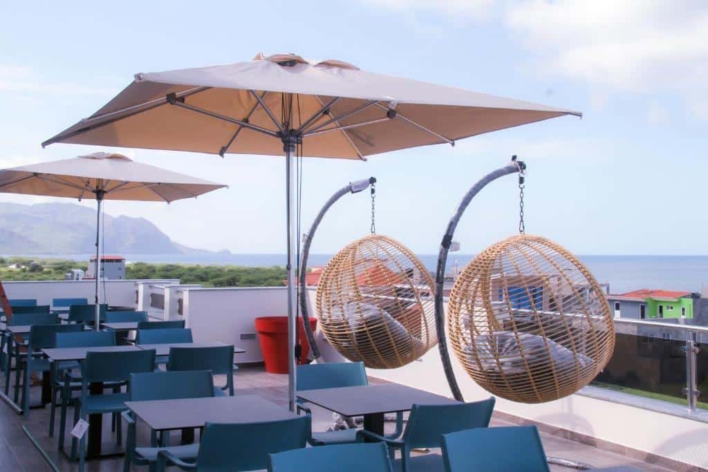 Terraço do Hotel Por Do Sol em Tarrafal que mostra várias mesas e cadeiras, dois guarda-sóis e dois balanços redondos de frente para a paisagem da cidade durante o dia. Imagem ilustrando post Hotéis em Cabo Verde.