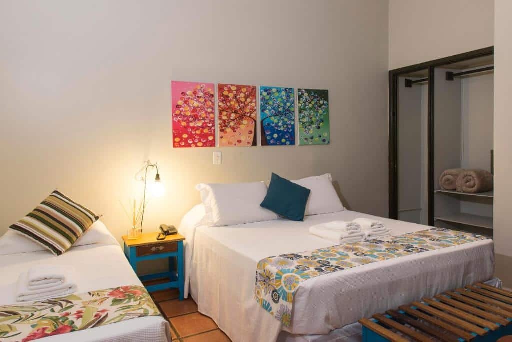 Quarto do Hotel Porto Di Mare. Do lado direito uma cama de solteiro, no meio uma mesa de apoio, do lado esquerdo uma cama de casal e na frente, um guarda-roupa, e na frente um banco.
