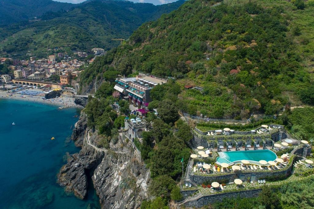 Imagem aérea do hotel mostrando a parte dos dormitórios e a piscina em cima da montanha ao lado do mar durante o dia, ilustrando post Hotéis em Cinque Terre.