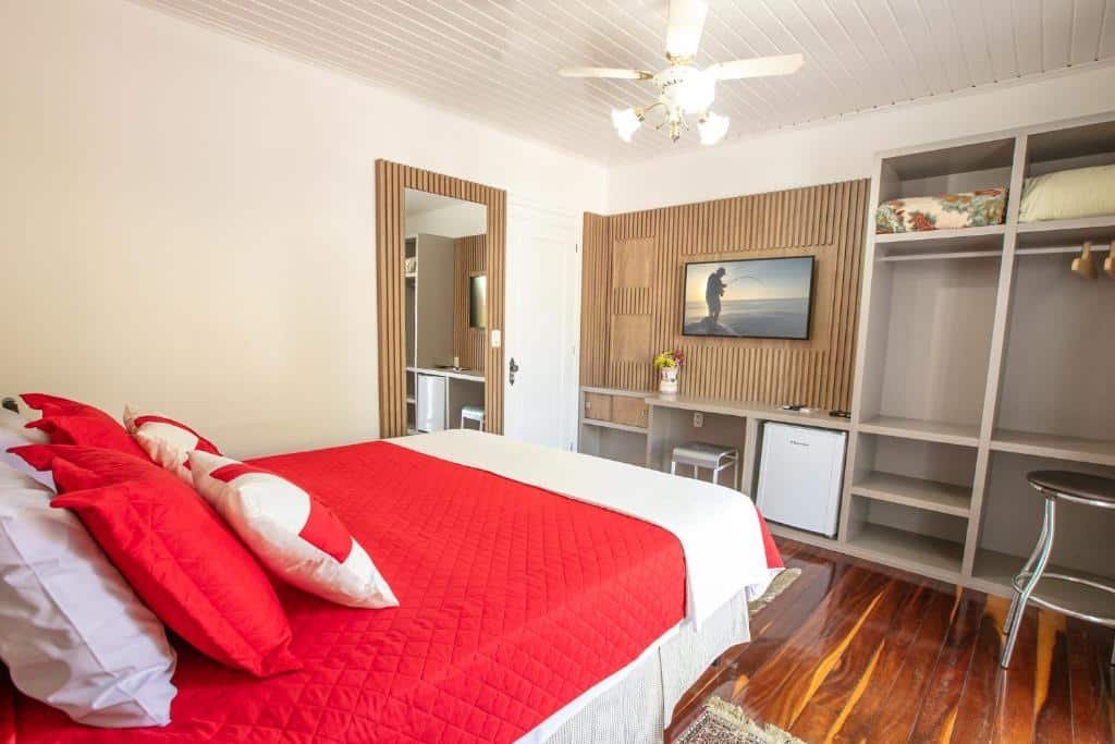 Quarto de casal no Hotel Pousada Casa Tasca, com frigobar, TV e armário, além de espelho de corpo inteiro e ventilador de teto. As roupas de cama são vermelhas