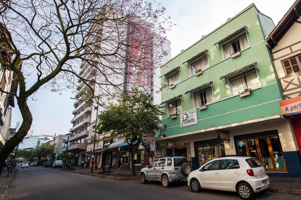 Rua com alguns carros andando e outros estacionados, uma uma árvore no lado esquerdo e, no lado direito, há prédios, sendo um deles um triplex verde escrito "HOTEL POUSADA XV" com letras azuis