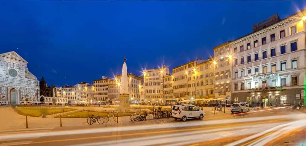 Início da noite em uma praça de Florença, com luzes acesas e vário prédios em volta, sendo um deles o Hotel Roma, que tem 4 andares (além do térreo)