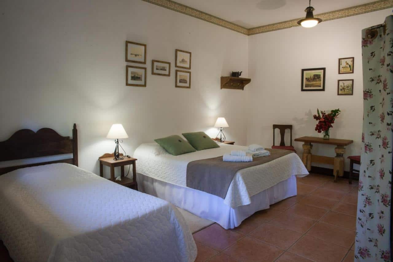 Um quarto com uma cama de casal e uma cama de solteiro, duas mesas de cabeceira com luminárias, quadros emoldurados na parede e uma pequena mesa com cadeiras no Hotel Solar dos Montes.