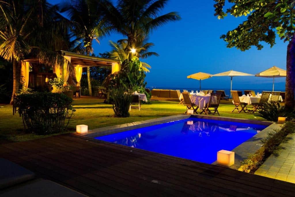 Piscina em Hotel Spa Nau Royal, com mesas e cadeiras no fim da tarde e início da noite de frente para o mar, muitas árvores e um bangalô do lado esquerdo, para representar pousadas em São Sebastião