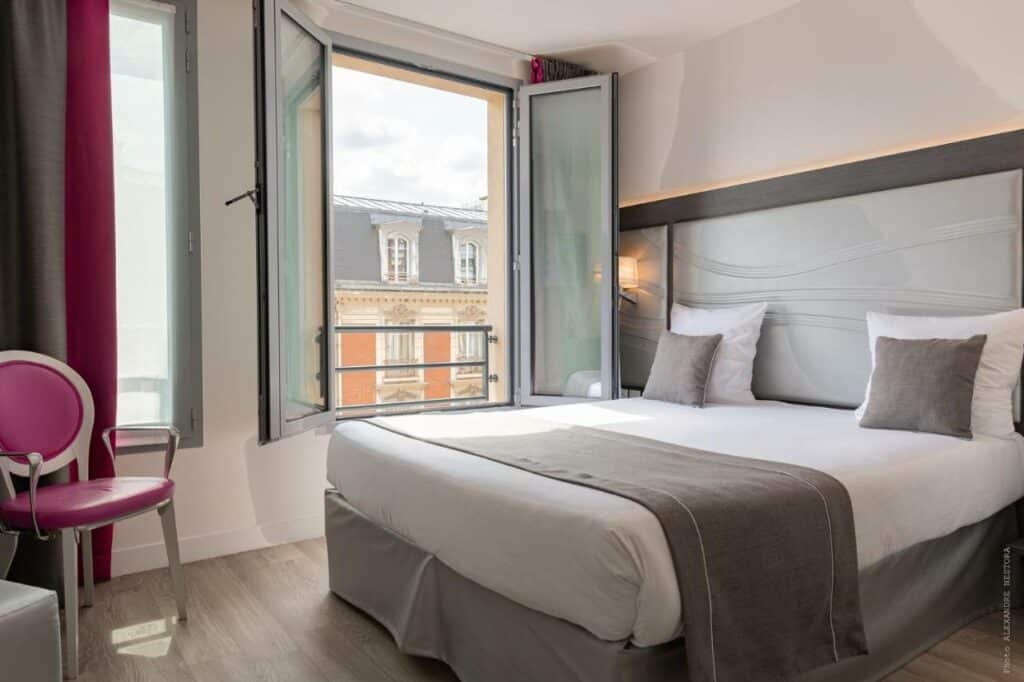 Quarto do Hôtel Versailles Chantiers, um dos hotéis perto do Palácio de Versalhes. Uma cama de casal está encostada na parede ao lado direito e conta com janelas com cortinas na lateral esquerda. Há também uma cadeira em frente à janela.