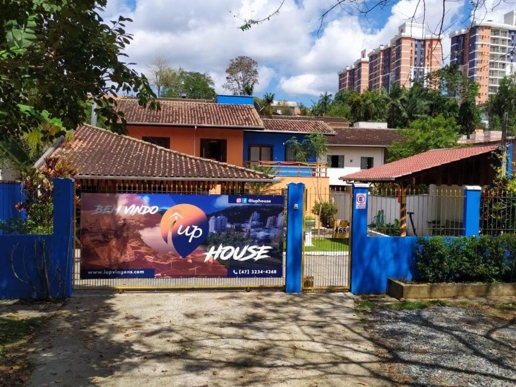 Frente da iUP HOUSE Hostel, com paredes azuis e uma placa grande escrito "Bem-vindo Up House - www.iupviagens.com - (47) 3234-4268"