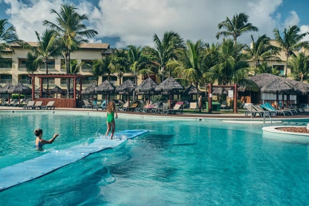 Duas pessoas aproveitam um dia ensolarado na piscina do Iberostar Waves Bahia - All Inclusive com palmeiras, espreguiçadeiras e cabanas de palha ao fundo.