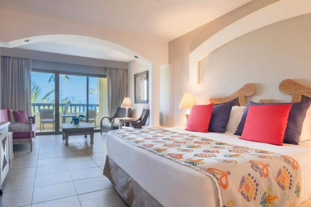 Quarto do Iberostar Waves Bahia - All Inclusive bem iluminado com uma cama grande, almofadas coloridas, área de estar e varanda com vista para as palmeiras e o oceano.