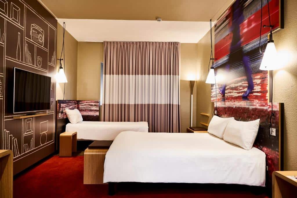 Quarto do Ibis Milano Centro com uma cama de casal, uma de solteiro, uma janela com cortinas, uma televisão e há algumas luminárias presas no teto