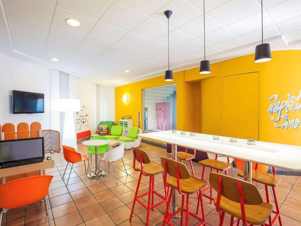 Parte do Ibis Styles Napoli Garibaldi com uma mesa com computador, uma tv na parede, algumas mesas diferentes com cadeiras espalhadas pelo ambiente e lustres pretos pendurados, ilustrando post Hotéis em Nápoles.