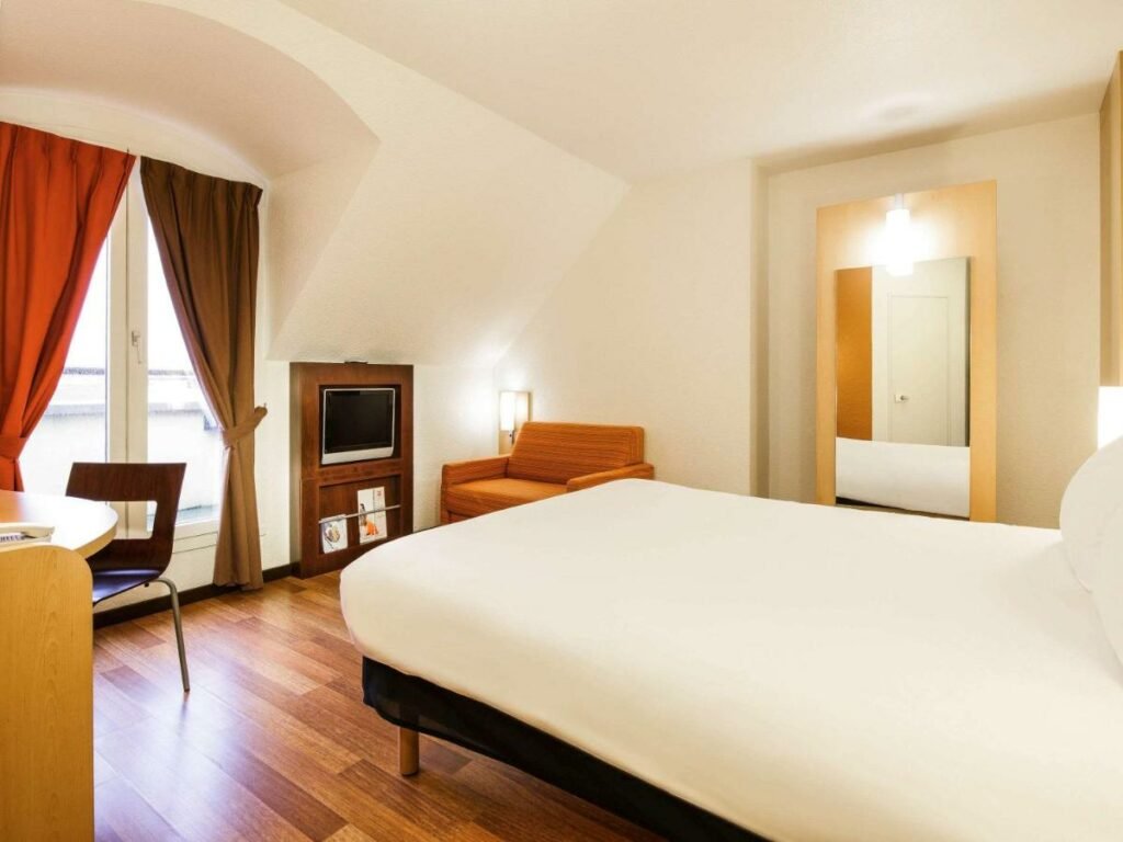 Quarto do ibis Versailles Château, um dos hotéis perto do Palácio de Versalhes. Uma cama de casal está encostada na parede ao lado direito e encara uma TV e uma poltrona na parede oposta. Ao lado esquerdo da cama há uma mesa e uma cadeira em frente à janela com cortinas. Já ao lado esquerdo há um espelho iluminado.