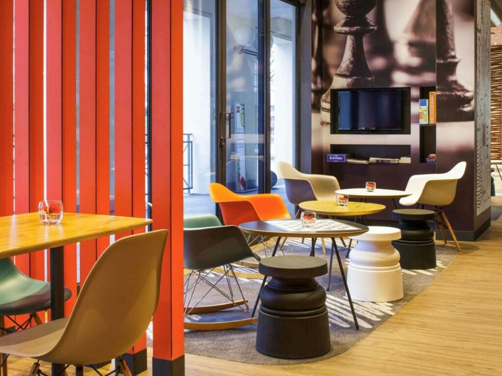 Área comum do ibis Versailles Château com mesinhas e cadeiras. Ao fundo há uma parede com jogos e TV. Três mesas ficam ao lado dessa TV, e outras são separadas por uma meia parede.