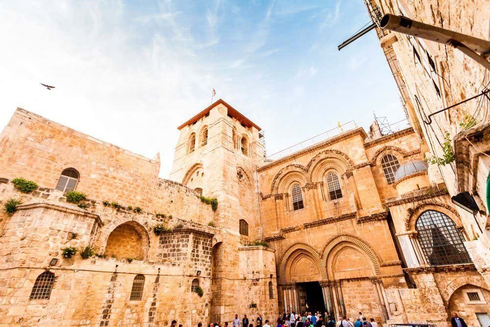 Um grupo de pessoas entra na Igreja do Santo Sepulcro, um edifício histórico de pedra com arcos e uma torre central, sob um céu azul em Jerusalém. Representa o que fazer em Israel.