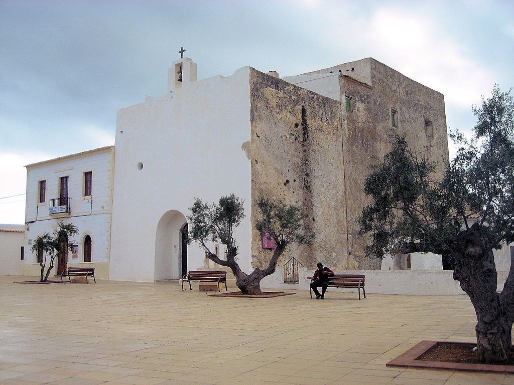 Uma pequena praça em Sant Francesc Xavier com bancos e oliveiras perto de uma igreja branca sob um céu nublado. Uma pessoa senta-se em um banco.