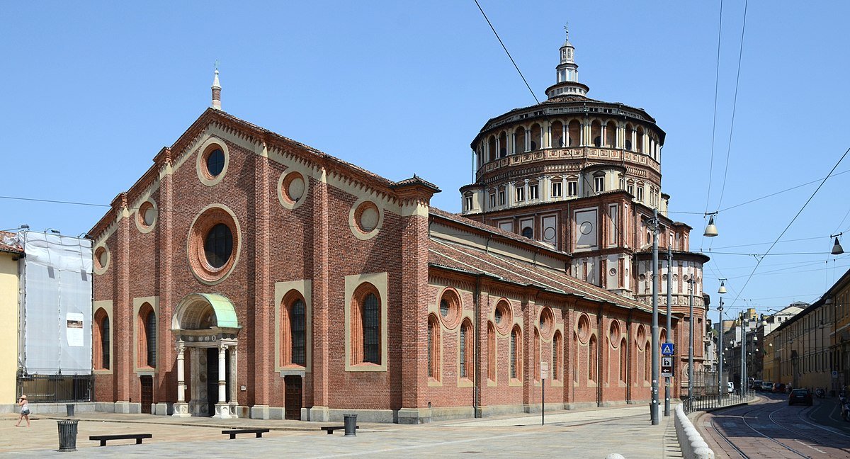 Vista de fora da Igreja de Santa Maria delle Grazie. O edifício é de tijolos vermelhos com fachada decorativa e uma grande cúpula de vários níveis ao fundo, situado em uma rua de paralelepípedos. Foto para o post de o que fazer em Milão.
