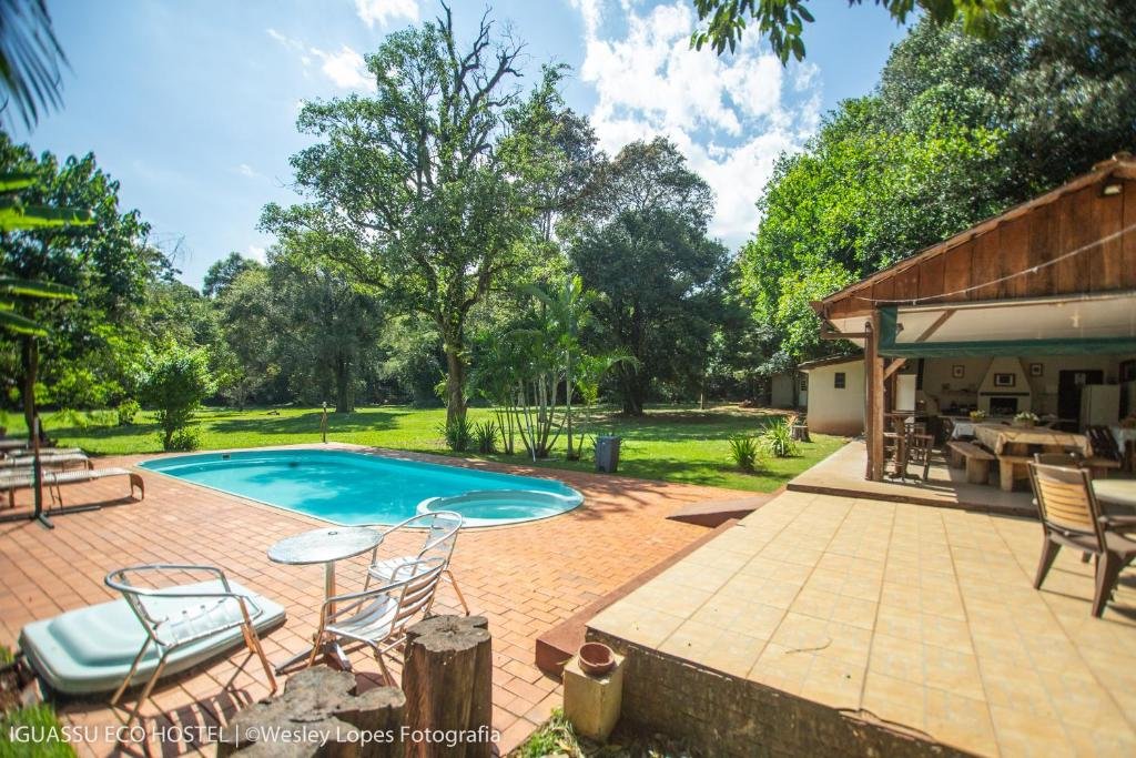 Parte do Iguassu Eco Hostel que mostra do lado esquerdo uma piscina ao ar livre com cadeiras, espreguiçadeiras e árvores verdes em volta. Do lado direito tem uma cobertura com mesas, cadeiras e outros móveis. Imagem ilustrando post Resorts em Foz do Iguaçu.