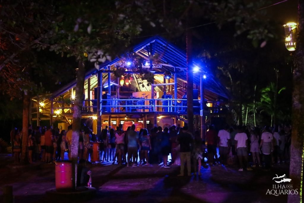 Foto da Ilha dos Aquários durante a noite em Porto Seguro. Há pessoas reunidas em frente à uma cabana iluminada. 