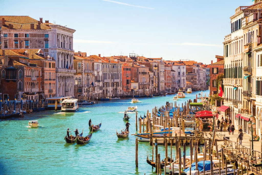 Vista do Grande Canal, em Veneza, com várias gôndolas e barcos pelo rio, além de outras atracadas, há diversas casinhas e prédios coloridos com muitas janelinhas ao estilo clássico italiano