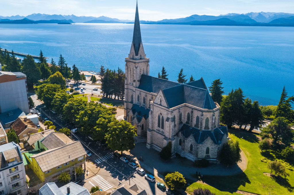 Vista de cima da Catedral de San Carlos de Bariloche, uma estrutura imponente com arquitetura alemã, em meio à grama e com o lago muito azul mais ao fundo