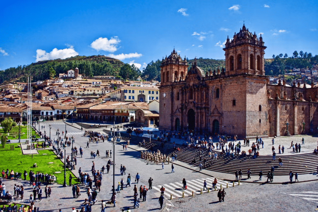 Vista de cima da Plaza de Armas, com praças de grama verdes e várias casinhas ao estilo colonial interiorano, no centro, há uma igreja bem grande com fachada de pedra e bem trabalhada em tons barrosos, ao longe, árvores verdes e céu azul