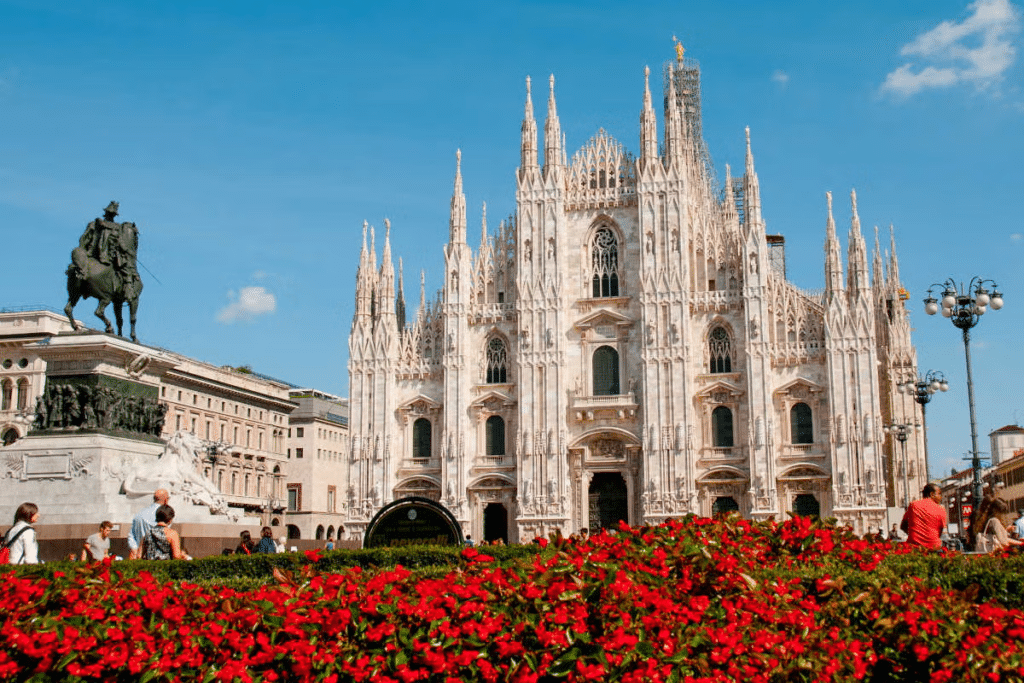 Vista do Duomo de Milão com muitas flores vermelhas à frente em um jardim, há uma escultura de um cavaleiro que aponto para a imponente catedral em estilo neogótico e fachada branca, com muitas torres pontudas e ricamente trabalhadas 