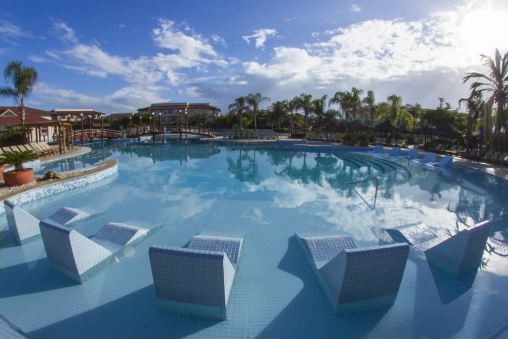 A imagem mostra uma ampla piscina ao ar livre no Grand Palladium Imbassaí Resort & Spa, cercada por cadeiras de praia e áreas de descanso. A água da piscina reflete o céu azul com algumas nuvens brancas, criando uma atmosfera serena e relaxante. Há cadeiras parcialmente submersas na água, permitindo que os hóspedes se refresquem enquanto relaxam. A vegetação ao redor inclui palmeiras e outras plantas tropicais, adicionando ao ambiente uma sensação de paraíso. Ao fundo, é possível ver parte da infraestrutura do resort, com edifícios e pontes que conectam diferentes áreas da piscina.