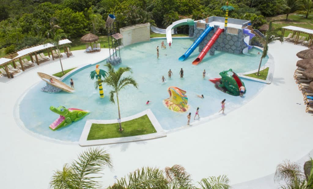 A imagem mostra um parque aquático infantil no Grand Palladium Imbassaí Resort & Spa, um dos melhores resorts all inclusive no Brasil. A piscina rasa está equipada com diversos brinquedos e tobogãs coloridos, proporcionando um ambiente divertido e seguro para as crianças. Os brinquedos têm formatos de animais e objetos, como um jacaré, uma rã e uma prancha de surfe. Palhoças com cadeiras ao redor da piscina oferecem áreas de sombra para os pais e responsáveis enquanto observam as crianças brincando. A vegetação tropical ao fundo adiciona uma sensação de natureza e tranquilidade ao ambiente.