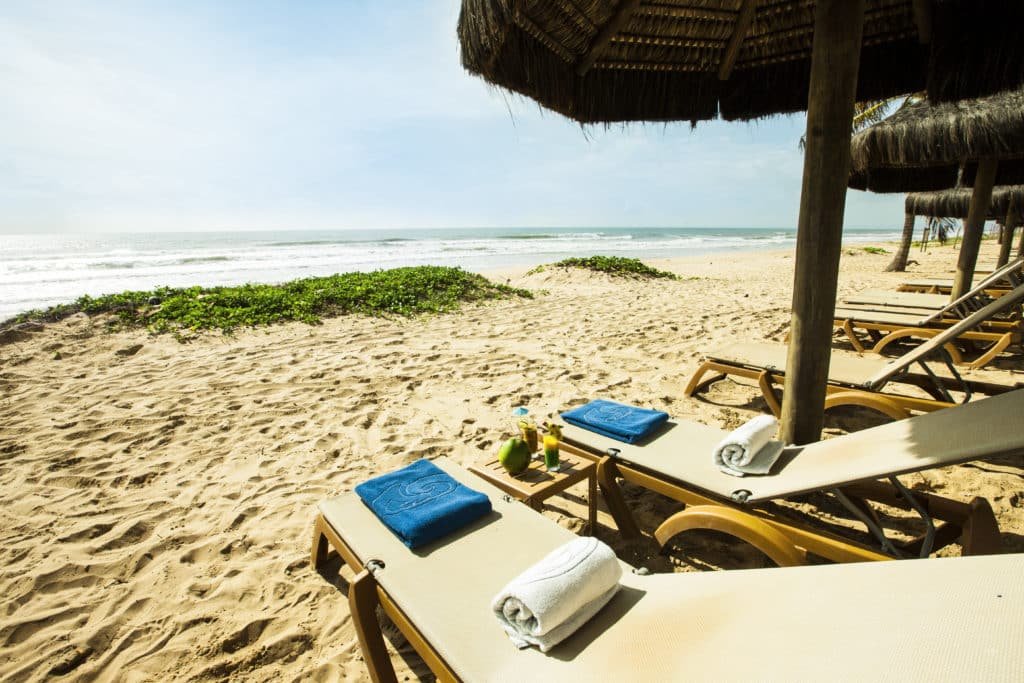 A imagem mostra uma praia serena e convidativa, com cadeiras de praia dispostas sob sombreiros de palha perto do Grand Palladium Imbassaí Resort & Spa, um dos melhores resorts all inclusive no Brasil. Toalhas e bebidas refrescantes, como água de coco, estão disponíveis nas espreguiçadeiras, criando um ambiente relaxante e confortável para os hóspedes aproveitarem o sol e a vista do mar. A areia é limpa e macia, e o mar ao fundo está calmo, com ondas suaves quebrando na praia.