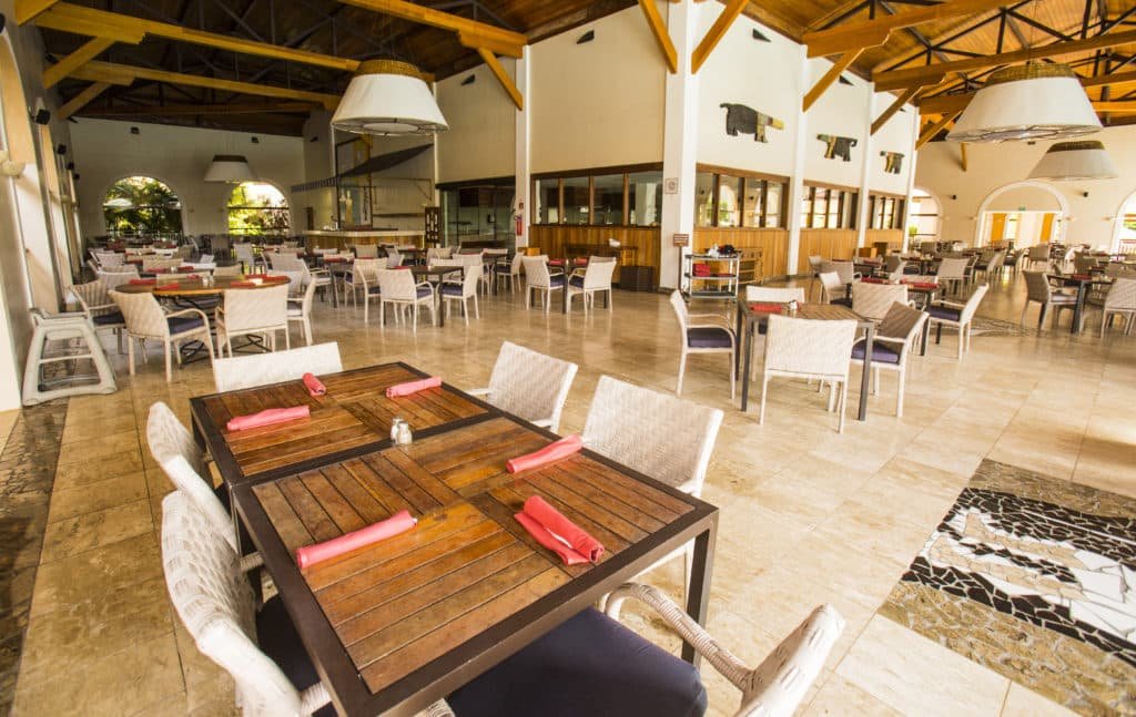 A imagem mostra um restaurante espaçoso e arejado no Grand Palladium Imbassaí Resort & Spa, decorado com mesas de madeira e cadeiras de vime. O teto alto com vigas de madeira expostas e luminárias grandes dão um toque rústico e acolhedor ao ambiente. As mesas estão organizadas com guardanapos vermelhos, prontos para receber os clientes. As janelas grandes permitem a entrada de muita luz natural, criando uma atmosfera agradável e iluminada.