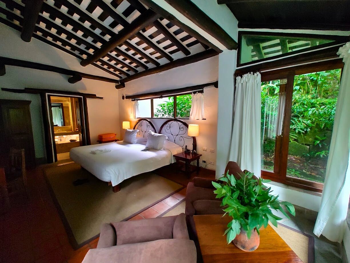 quarto do Inkaterra Machu Picchu Pueblo Hotel, no Peru, com uma cama de casal ao centro, uma área de estar com duas poltronas no canto direito inferior, ao lado de uma ampla janela com vista para a vegetação. Na outra ponta do quarto é possível enxergar a porta aberta para o banheiro da suíte