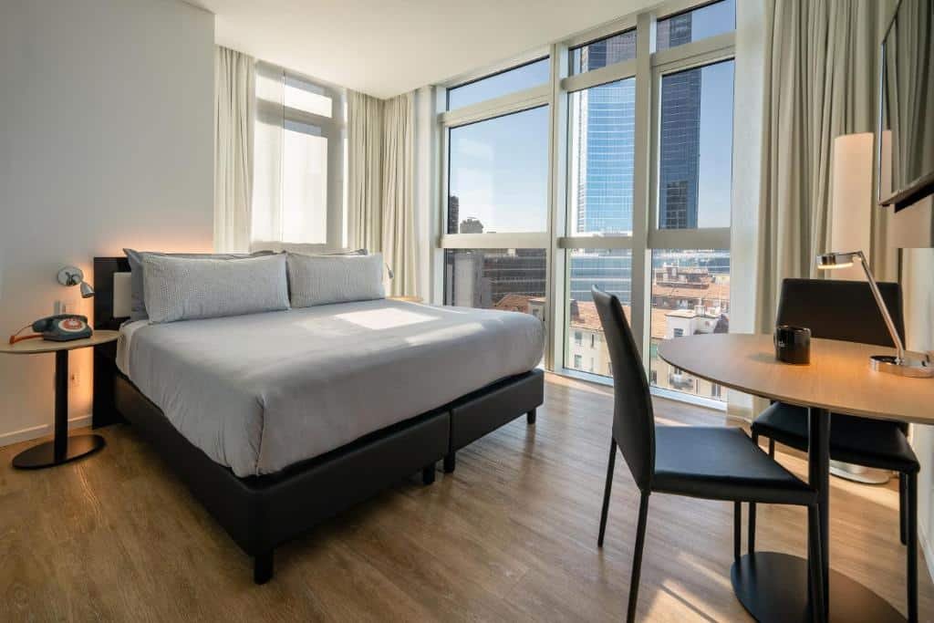 Quarto do INNSiDE by Meliá Milano Torre GalFa com uma janela panorâmica com vista para a cidade, há uma cama de casal, uma televisão e uma mesinha redonda com duas cadeiras