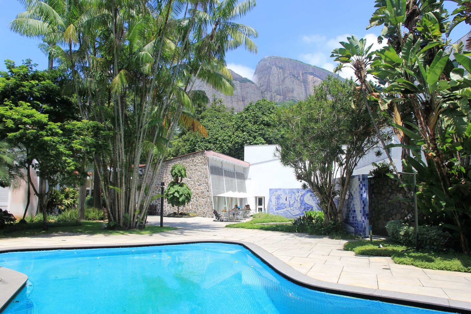 Uma piscina é cercada por árvores e plantas tropicais. Prédios com paredes brancas estão ao fundo. Uma montanha é visível.