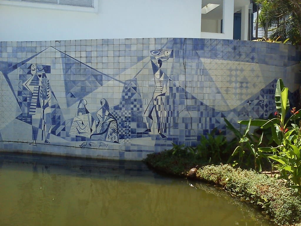 Mural de azulejos azuis representando figuras abstratas ao lado de um lago, com vegetação em primeiro plano e um edifício branco ao fundo. Imagem para ilustrar o post sobre museus no Rio de Janeiro.