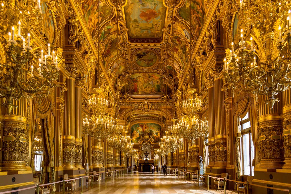 Hall ornamentado no Palácio de Versalhes com teto alto adornado com afrescos, detalhes dourados, lustres e janelas altas. Representa um grande design arquitetônico.