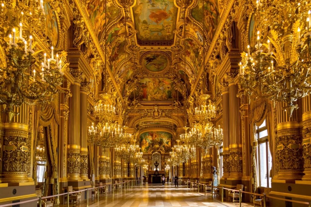 Um salão grandioso e ornamentado com decorações douradas, lustres e tetos pintados, criando uma atmosfera opulenta. Esse é o interior de uma sala no Palácio de Versalhes, um dos passeios em Paris.
