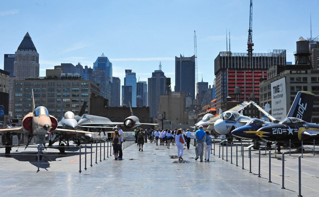 Pessoas caminhando no convés do Intrepid Sea, Air & Space Museum exibindo vários aviões antigos, com o horizonte da cidade ao fundo.