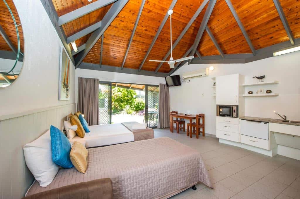 Quarto espaçoso com duas camas, teto de madeira, cozinha compacta, mesa de jantar, TV montada na parede e vista para a área verde do lado de fora.