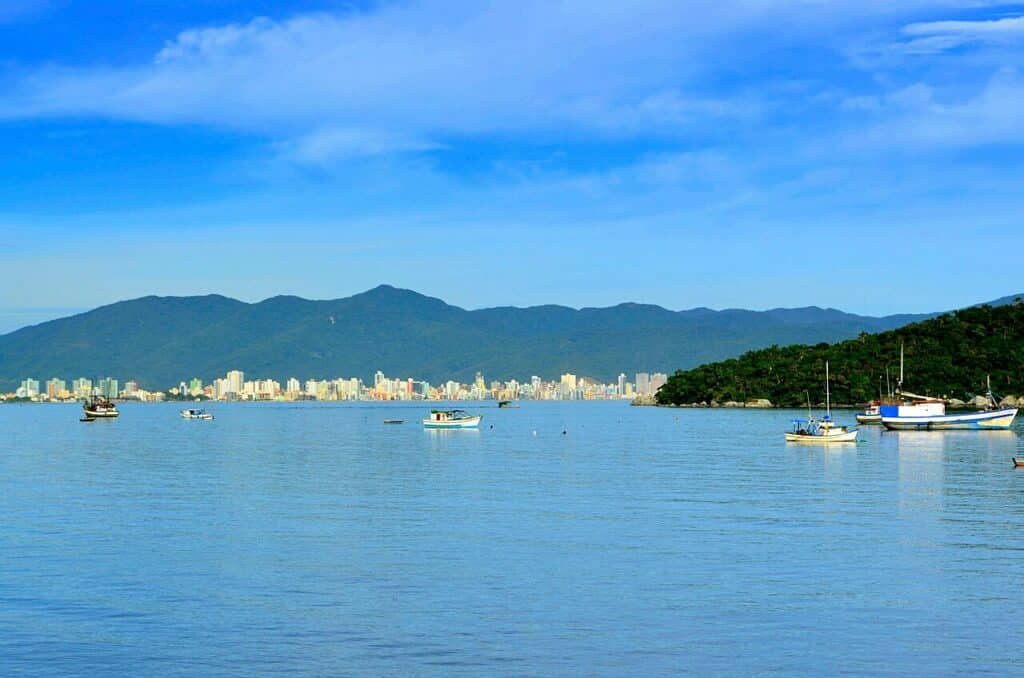 Itapema com montanhas ao fundo, uma orla com prédios e barcos no mar azul