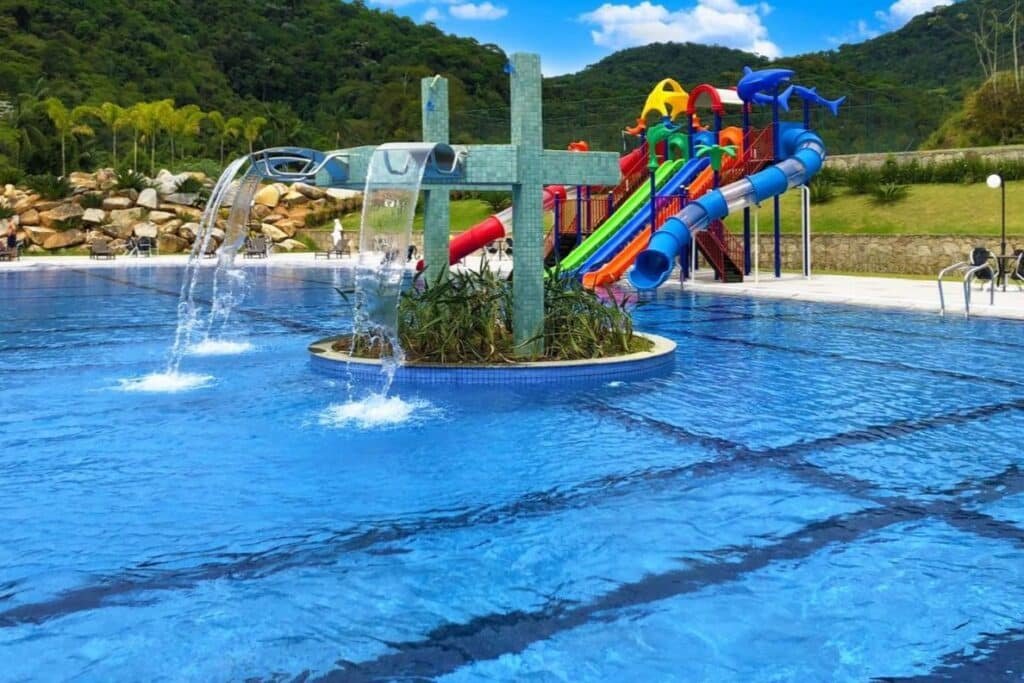 Uma piscina do Itatiaia Resort & Eventos, um dos hotéis fazenda no Rio de Janeiro, conta com uma área de toboágua com escorregadores coloridos e uma fonte, cercada por árvores e colinas ao fundo.