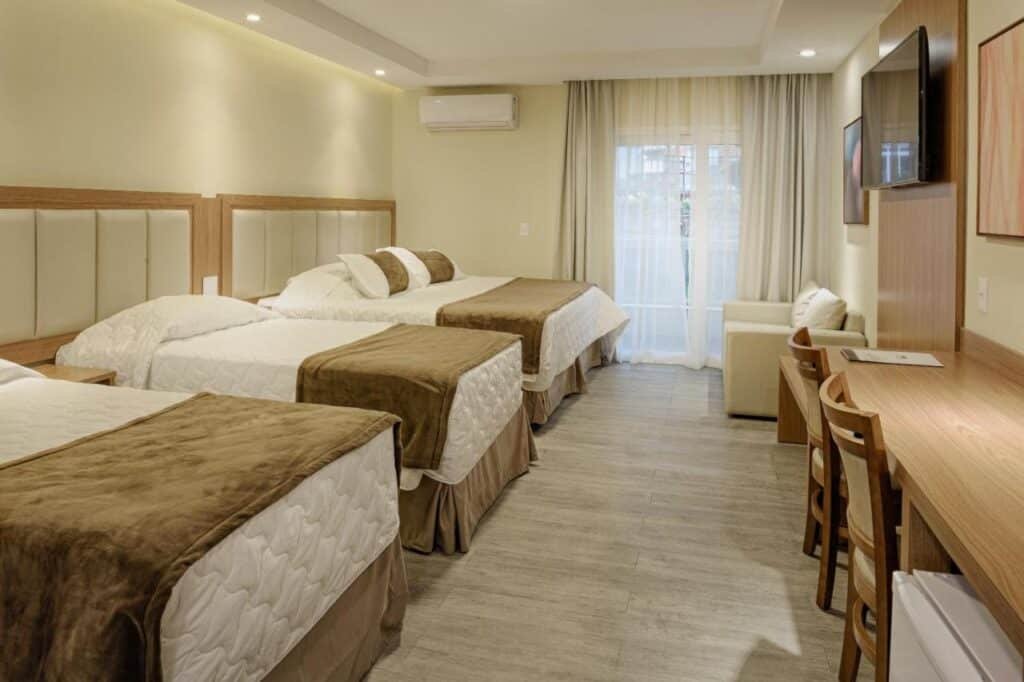 Quarto do Itatiaia Resort & Eventos com três camas, roupa de cama bege e marrom, uma escrivaninha de madeira, cadeiras, TV montada na parede e uma janela com cortinas transparentes.