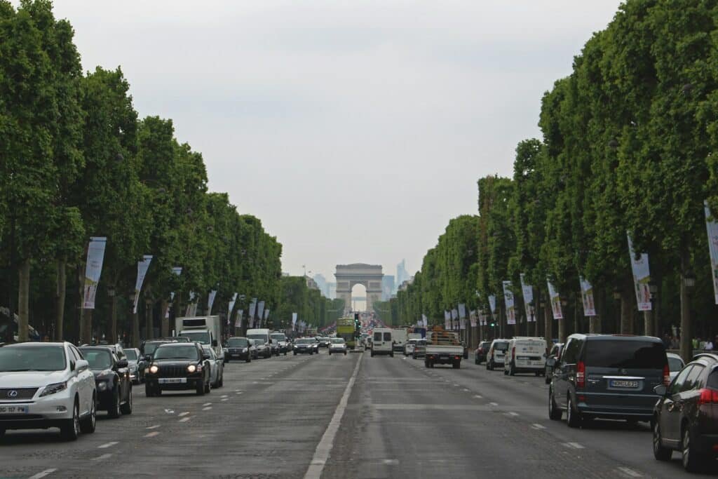 A imagem mostra uma ampla avenida ladeada por árvores verdes, com carros circulando em ambos os sentidos. Ao fundo, destaca-se o Arco do Triunfo, um famoso monumento parisiense. A avenida é a Champs-Élysées, uma das mais conhecidas de Paris, e está coberta por um céu nublado, criando uma atmosfera tranquila e típica de dias em que o sol está encoberto. Banners publicitários estão pendurados ao longo da avenida, adicionando cor ao cenário. Esse é um dos pontos turísticos de Paris.