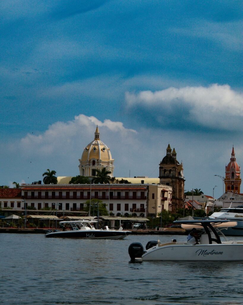 A imagem mostra a vista do mar em direção ao centro histórico de Cartagena, destacando a Igreja de Santo Domingo e a Catedral de Santa Catalina de Alejandría. No primeiro plano, há barcos ancorados na água, incluindo um com o nome "Malú" e outro chamado "Martran". Ao fundo, a cúpula amarela e branca da catedral e as torres das igrejas se erguem contra o céu azul com algumas nuvens. A arquitetura colonial dos edifícios próximos à água complementa a paisagem, criando uma cena pitoresca e histórica. - Foto: Jaime A Vega via Pexels