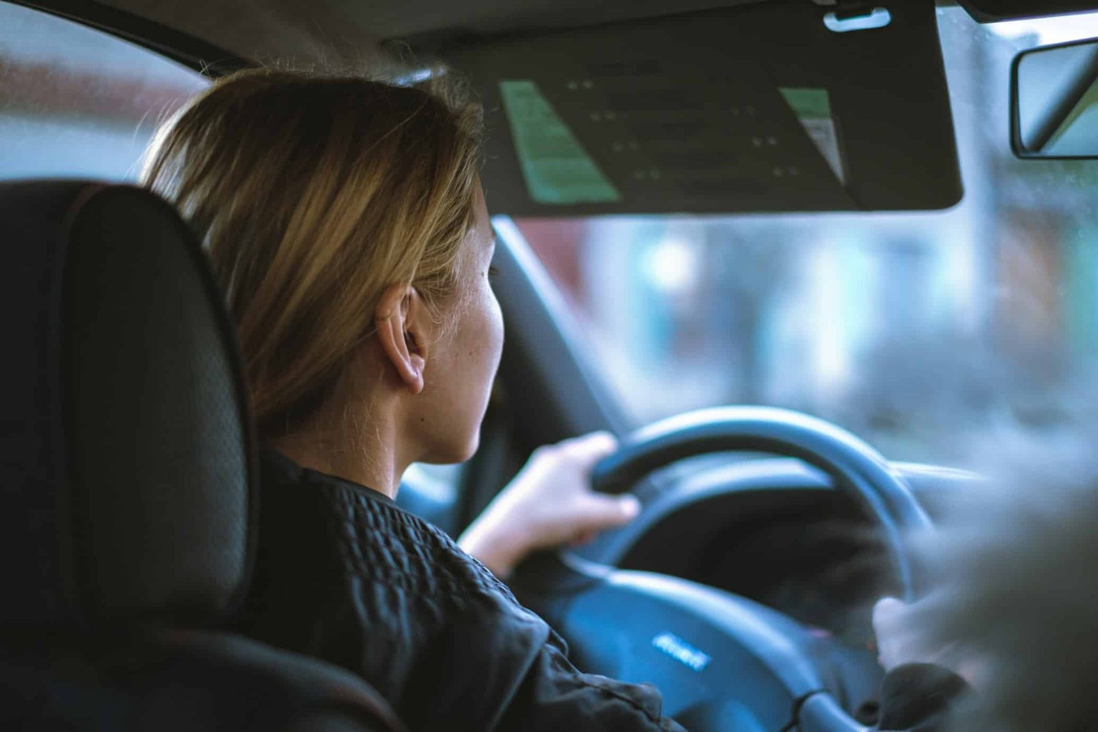 Pessoa com cabelo longo dirigindo um carro, vista do banco de trás. O foco está no motorista e no volante.