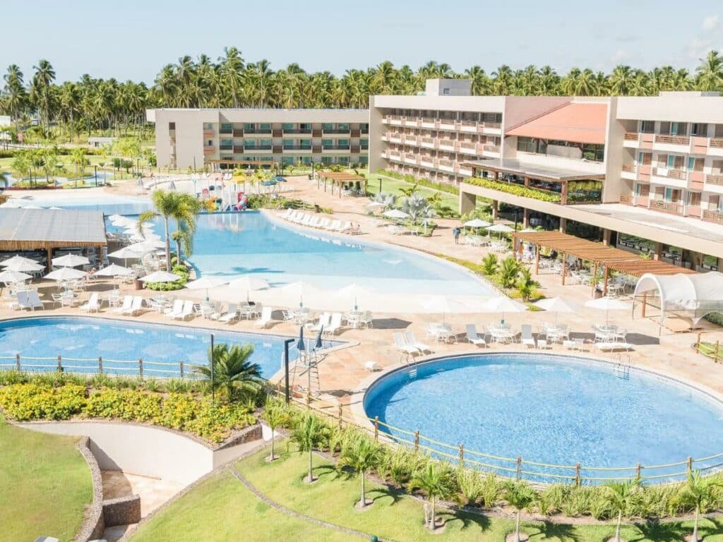 A imagem mostra uma visão aérea do Japaratinga Lounge Resort, destacando a ampla área de lazer com diversas piscinas de formatos e tamanhos diferentes. No centro, uma grande piscina principal é cercada por espreguiçadeiras brancas e guarda-sóis, proporcionando um ambiente relaxante para os hóspedes. À esquerda, uma piscina menor com cobertura de palha e mais espreguiçadeiras. Ao fundo, pode-se ver o prédio principal do resort, com várias varandas voltadas para a área da piscina. O cenário é complementado por palmeiras e vegetação exuberante, criando uma atmosfera tropical e convidativa.